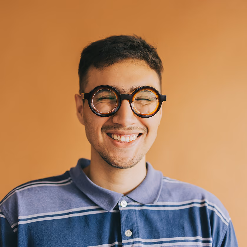 Young man smiling with eyes closed wearing round tortoiseshell glasses and a striped polo shirt.