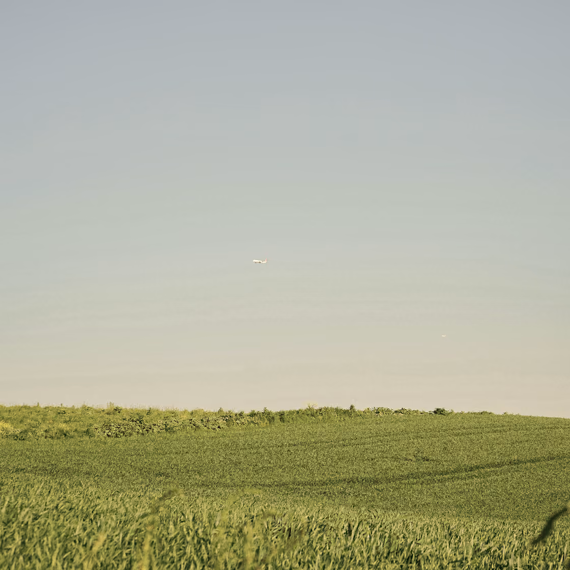 Small airplane flying over green grassy fields under a pale blue sky.